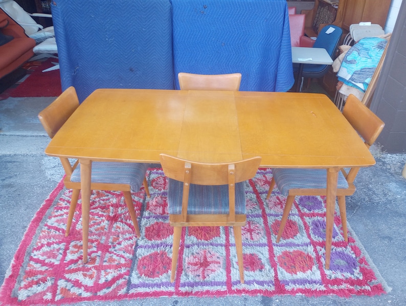 May include: A set of four mid-century modern dining chairs with a light wood frame and gray fabric seats. The chairs are arranged around a rectangular table with a light wood top. The table and chairs are on a red and purple rug.