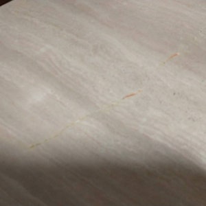 May include: A close-up of a light-colored, textured surface with subtle veining and streaks. The surface has a neutral color palette with hints of beige, cream, and light pink, creating a natural, organic appearance. The surface appears to be made of stone.
