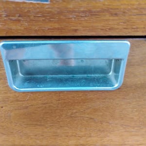 May include: A rectangular, recessed metal drawer pull with a polished silver finish, set into a smooth, medium-brown wooden surface with a visible grain pattern. The handle features a clean, horizontal design with rounded corners and a deep inset.