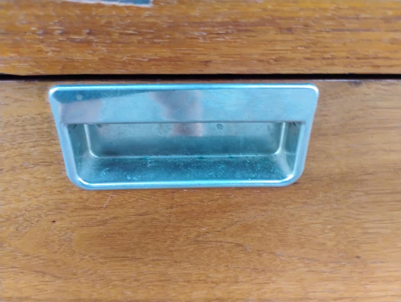 May include: A rectangular, recessed metal drawer pull with a polished silver finish, set into a smooth, medium-brown wooden surface with a visible grain pattern. The handle features a clean, horizontal design with rounded corners and a deep inset.