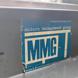 May include: A dark grey metal surface featuring a rectangular blue sticker with white text. The sticker displays the text "modern management group", "MMG", "designed by george nelson", and "herman miller furniture company" with visible wear.