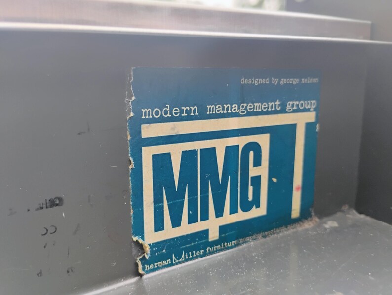 May include: A dark grey metal surface featuring a rectangular blue sticker with white text. The sticker displays the text "modern management group", "MMG", "designed by george nelson", and "herman miller furniture company" with visible wear.