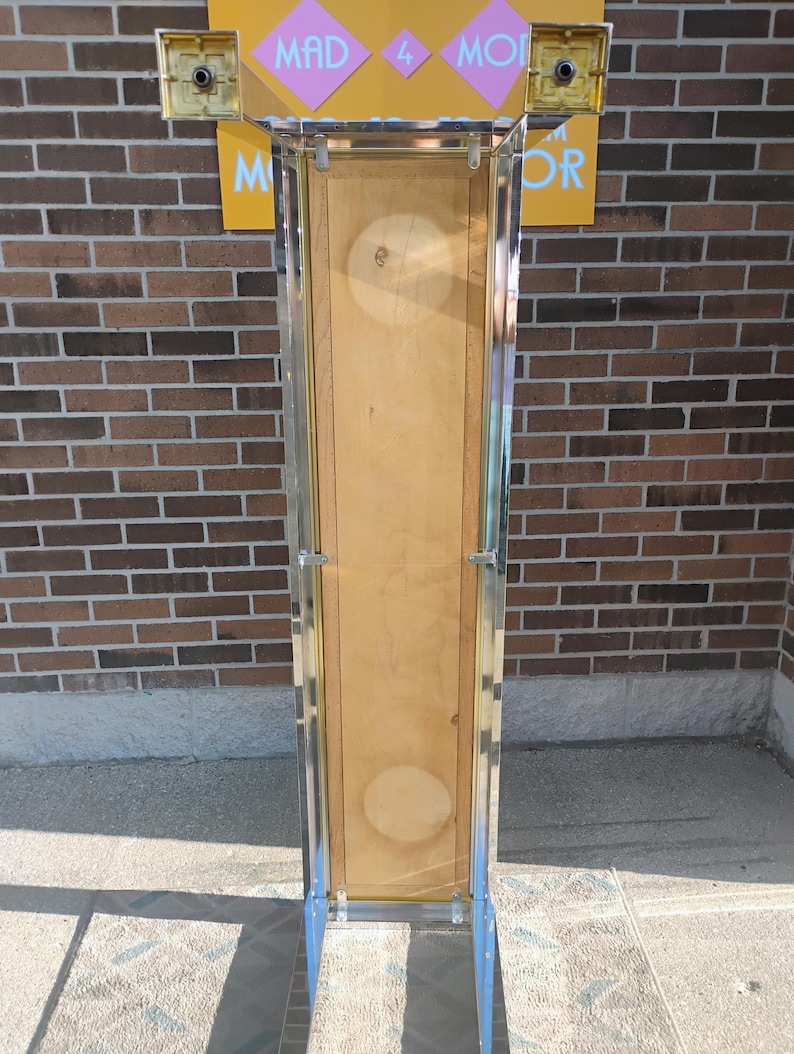 K&ouml;nnte beinhalten: Eine hohe, rechteckige Vitrine mit einer Holzr&uuml;ckwand und einer transparenten Vorderseite. Der Rahmen ist aus silberfarbenem Metall. &Uuml;ber der Vitrine befindet sich ein Schild mit der Aufschrift "MAD 4 MOR" in rosa und wei&szlig;er Schrift auf gelbem Hintergrund. Die Vitrine steht auf einer Betonoberfl&auml;che.