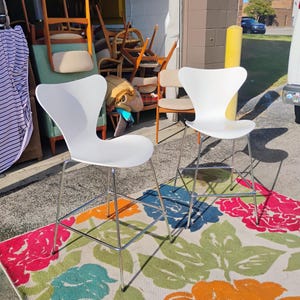 K&ouml;nnte beinhalten: Zwei wei&szlig;e, moderne Barhocker mit Chrombeinen stehen auf einem bunten Blumengemusterte Teppich. Der Teppich zeigt gro&szlig;e Blumen in Rosa, Blau, Orange und Gr&uuml;n. Im Hintergrund sind weitere Vintage-St&uuml;hle zu sehen.