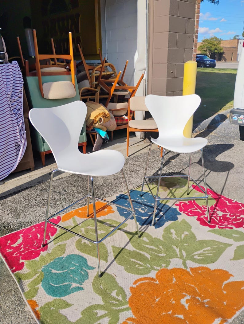 K&ouml;nnte beinhalten: Zwei wei&szlig;e, moderne Barhocker mit Chrombeinen stehen auf einem bunten Blumengemusterte Teppich. Der Teppich zeigt gro&szlig;e Blumen in Rosa, Blau, Orange und Gr&uuml;n. Im Hintergrund sind weitere Vintage-St&uuml;hle zu sehen.