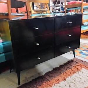 May include: A black six-drawer dresser with silver-toned drawer pulls. The dresser has a glossy finish and tapered legs. The furniture piece is in a room with other furniture and a patterned rug.