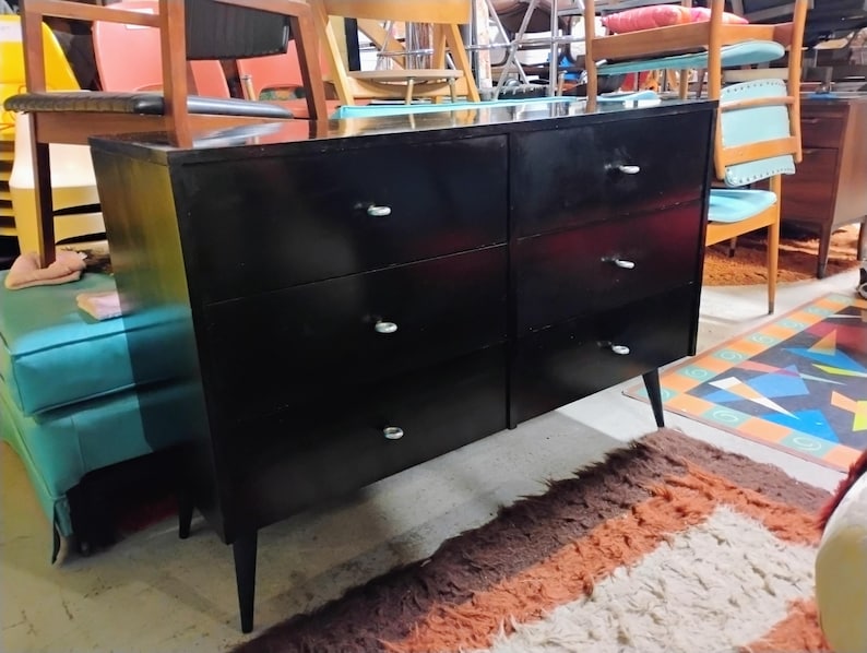 May include: A black six-drawer dresser with silver-toned drawer pulls. The dresser has a glossy finish and tapered legs. The furniture piece is in a room with other furniture and a patterned rug.