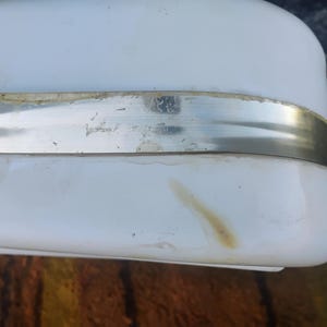 May include: A white rectangular object with rounded corners and a horizontal metallic band across the center. The surface features a visible light brown stain near the bottom edge and some residue along the top of the metal strip.