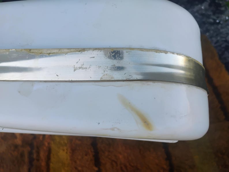 May include: A white rectangular object with rounded corners and a horizontal metallic band across the center. The surface features a visible light brown stain near the bottom edge and some residue along the top of the metal strip.