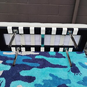 May include: A black and white bench with a white cushioned top and chrome legs. The bench has a rectangular frame with white straps and is placed on a blue, white, and navy camouflage patterned rug.