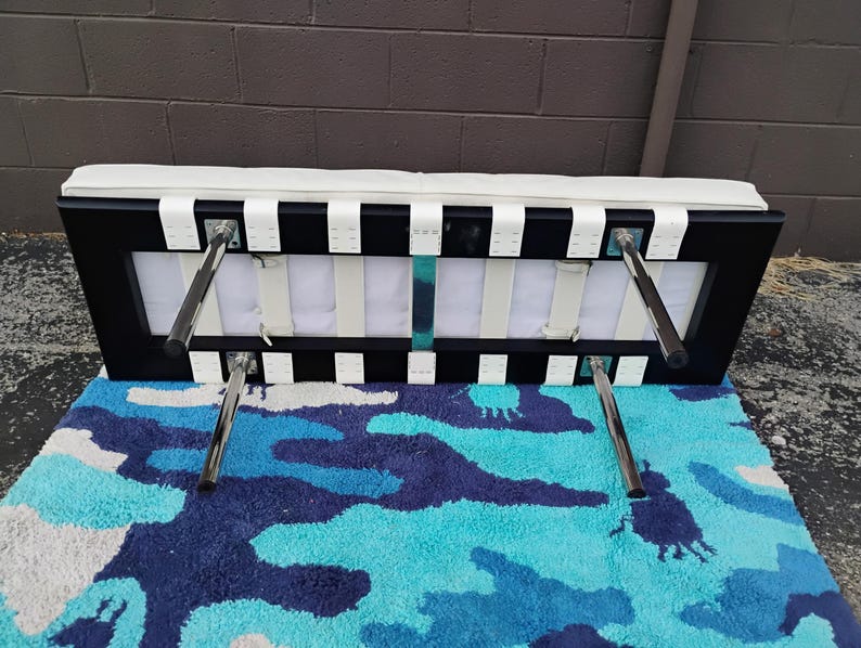 May include: A black and white bench with a white cushioned top and chrome legs. The bench has a rectangular frame with white straps and is placed on a blue, white, and navy camouflage patterned rug.