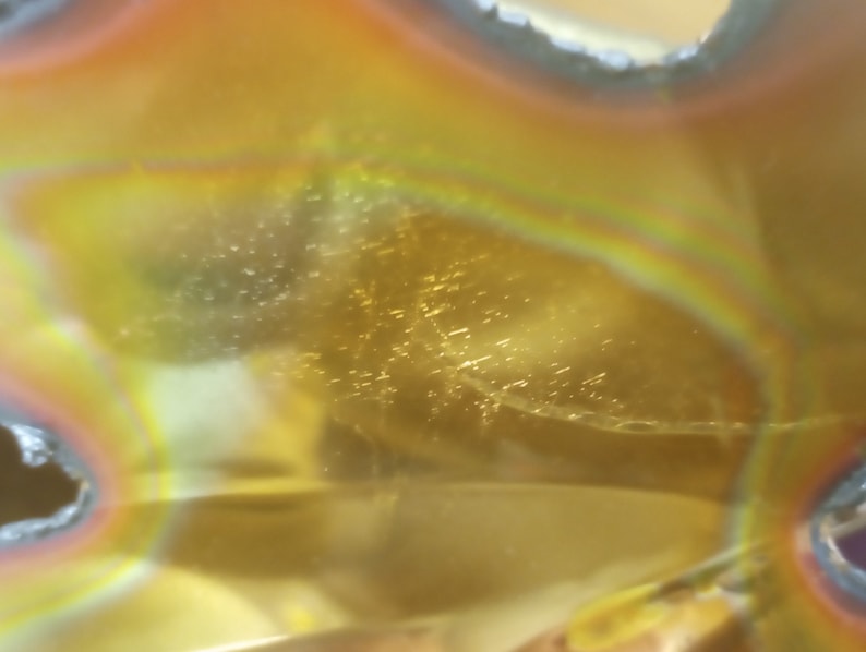 Puede incluir: Primer plano de un objeto de vidrio amarillo, naranja e iridiscente con una superficie texturizada. El vidrio tiene una forma circular con un agujero en el centro.