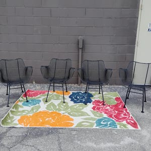 May include: Four black metal chairs with a wire mesh design are arranged around a colorful floral rug. The chairs have a simple, modern design and are perfect for outdoor seating.