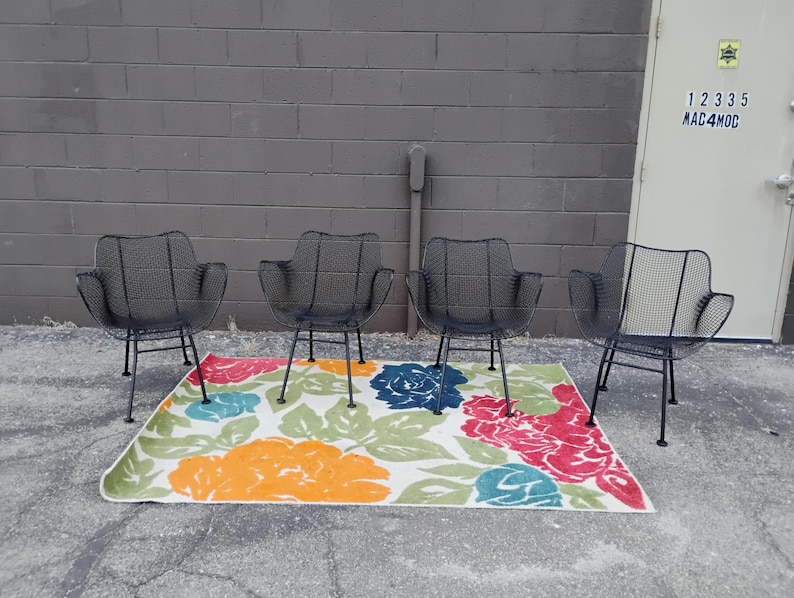 May include: Four black metal chairs with a wire mesh design are arranged around a colorful floral rug. The chairs have a simple, modern design and are perfect for outdoor seating.