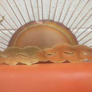 May include: A copper and brass wall sculpture depicting a sunset with a large, round, orange sun and three clouds. The sun has a rainbow-like pattern in the centre. The clouds are made of brass and have a wavy, organic shape.