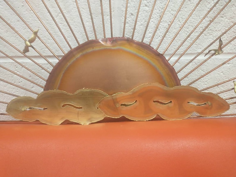 May include: A copper and brass wall sculpture depicting a sunset with a large, round, orange sun and three clouds. The sun has a rainbow-like pattern in the centre. The clouds are made of brass and have a wavy, organic shape.