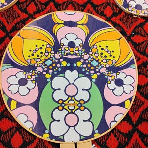 May include: Circular embroidery hoops with a retro floral design. The fabric features a pattern of large white, pink, yellow, and orange flowers on a dark blue background. The hoops are set against a red and black patterned backdrop.