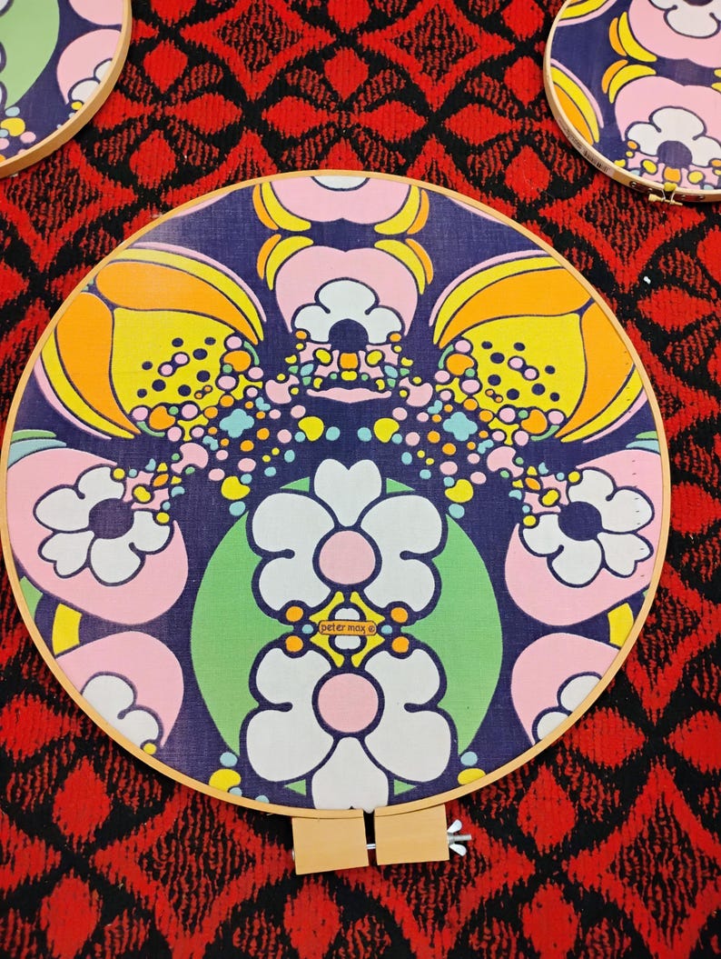 May include: Circular embroidery hoops with a retro floral design. The fabric features a pattern of large white, pink, yellow, and orange flowers on a dark blue background. The hoops are set against a red and black patterned backdrop.