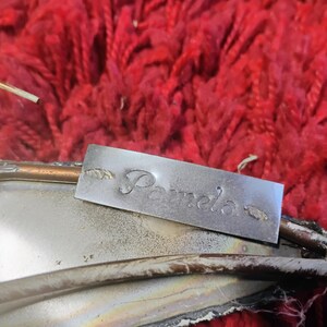 Pode incluir: Uma etiqueta de metal prateado com o nome "Pamela" gravado. A etiqueta &eacute; retangular e tem desenhos decorativos em forma de folha em cada lado do nome. A etiqueta est&aacute; colocada sobre um objeto prateado com uma borda cor de cobre, contra um fundo vermelho texturizado.