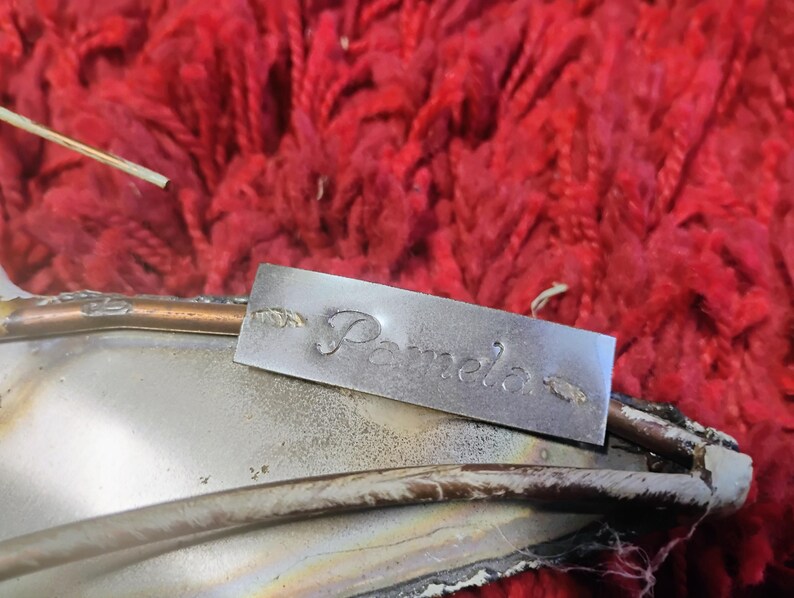 Pode incluir: Uma etiqueta de metal prateado com o nome "Pamela" gravado. A etiqueta &eacute; retangular e tem desenhos decorativos em forma de folha em cada lado do nome. A etiqueta est&aacute; colocada sobre um objeto prateado com uma borda cor de cobre, contra um fundo vermelho texturizado.
