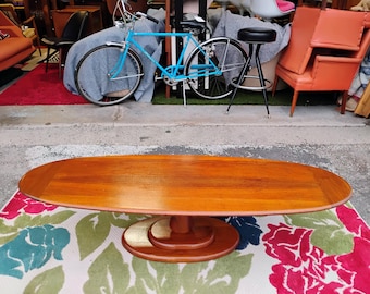 May include: A mid-century modern oval-shaped coffee table with a wooden top and a pedestal base. The table is made of teak wood and has a natural finish. The table is sitting on a floral rug.