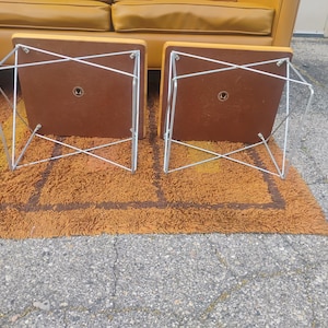 May include: Two square side tables with mustard yellow tops and brown undersides, featuring silver-toned metal wire frames in a crisscross pattern. The tables are positioned upside down, revealing the flat brown bases and the metal hardware.