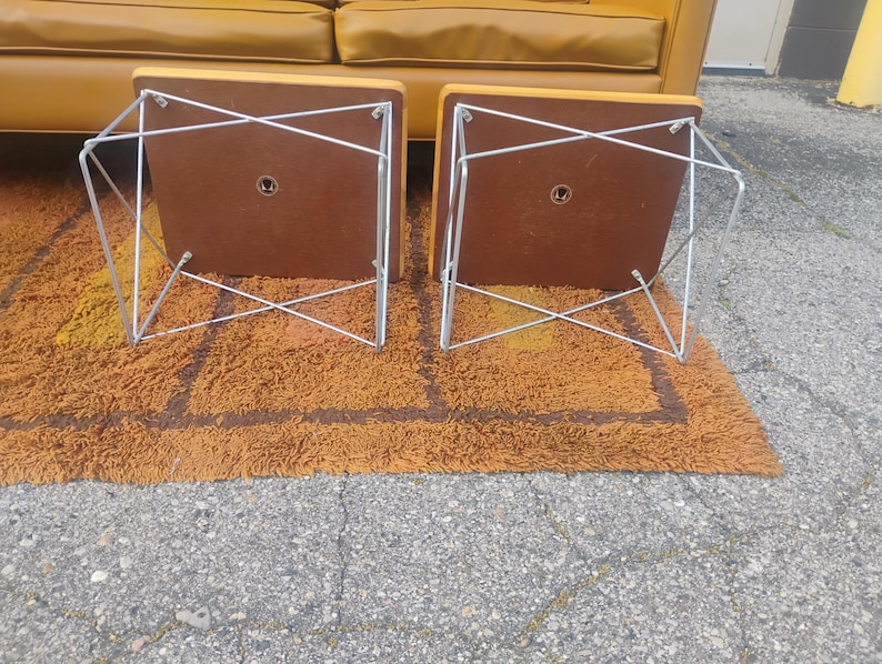 May include: Two square side tables with mustard yellow tops and brown undersides, featuring silver-toned metal wire frames in a crisscross pattern. The tables are positioned upside down, revealing the flat brown bases and the metal hardware.