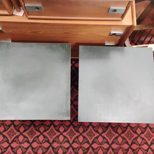 May include: Two square, dark gray tabletops with a smooth, matte finish. The surfaces are flat and feature a uniform, solid color. These square panels are positioned side-by-side, displaying a clean, geometric design with sharp, straight edges.