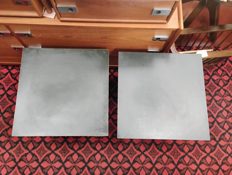 May include: Two square, dark gray tabletops with a smooth, matte finish. The surfaces are flat and feature a uniform, solid color. These square panels are positioned side-by-side, displaying a clean, geometric design with sharp, straight edges.