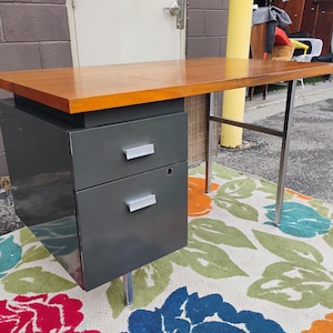 May include: A desk featuring a rectangular wooden top with a warm brown finish. The base consists of a dark grey metal cabinet with two stacked drawers, each equipped with a silver handle, supported by thin, silver-toned metal legs.