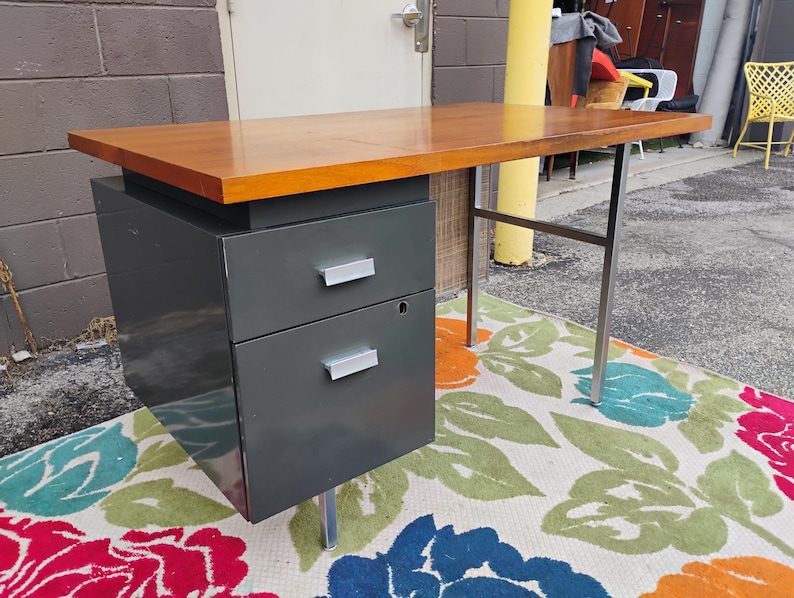 May include: A desk featuring a rectangular wooden top with a warm brown finish. The base consists of a dark grey metal cabinet with two stacked drawers, each equipped with a silver handle, supported by thin, silver-toned metal legs.