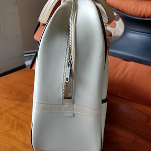 Pode incluir: Bolsa de viagem de vinil branco com acabamento preto e fecho de correr. A bolsa tem uma al&ccedil;a superior e uma al&ccedil;a de ombro.