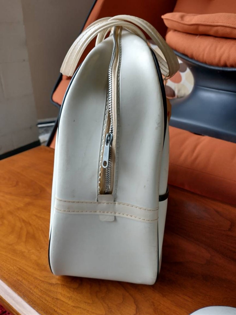 Pode incluir: Bolsa de viagem de vinil branco com acabamento preto e fecho de correr. A bolsa tem uma al&ccedil;a superior e uma al&ccedil;a de ombro.