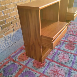 May include: Two wooden nightstands with a warm, natural finish. Each nightstand features a drawer and an open shelf. The nightstands are placed on a colorful, patterned rug, with a brick wall in the background.