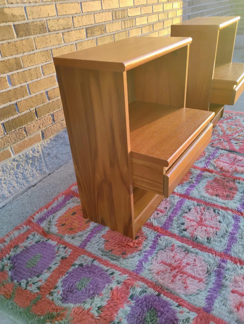 May include: Two wooden nightstands with a warm, natural finish. Each nightstand features a drawer and an open shelf. The nightstands are placed on a colorful, patterned rug, with a brick wall in the background.
