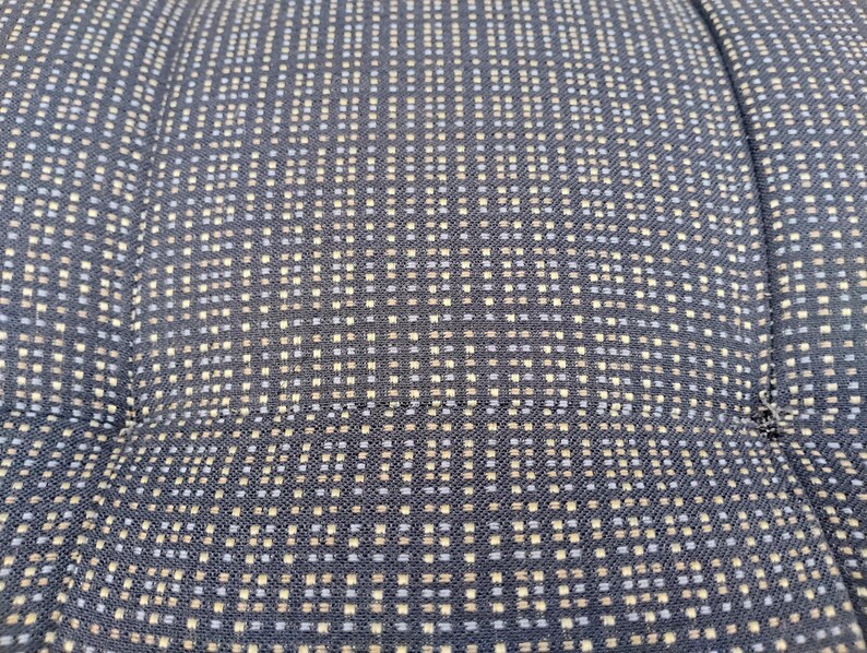 K&ouml;nnte beinhalten: Ein dunkelblauer Stoff mit einem sich wiederholenden Gittermuster aus kleinen hellblauen und beigen quadratischen Punkten. Das Textil weist eine sichtbare Webstruktur auf, wobei vertikale und horizontale N&auml;hte eine gesteppte Optik erzeugen.