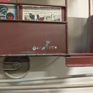 May include: A red metal model of a train car with a white inscription that reads "c.c. de 1996". The train car has a metal frame and a red body. The train car has two wheels on the bottom.