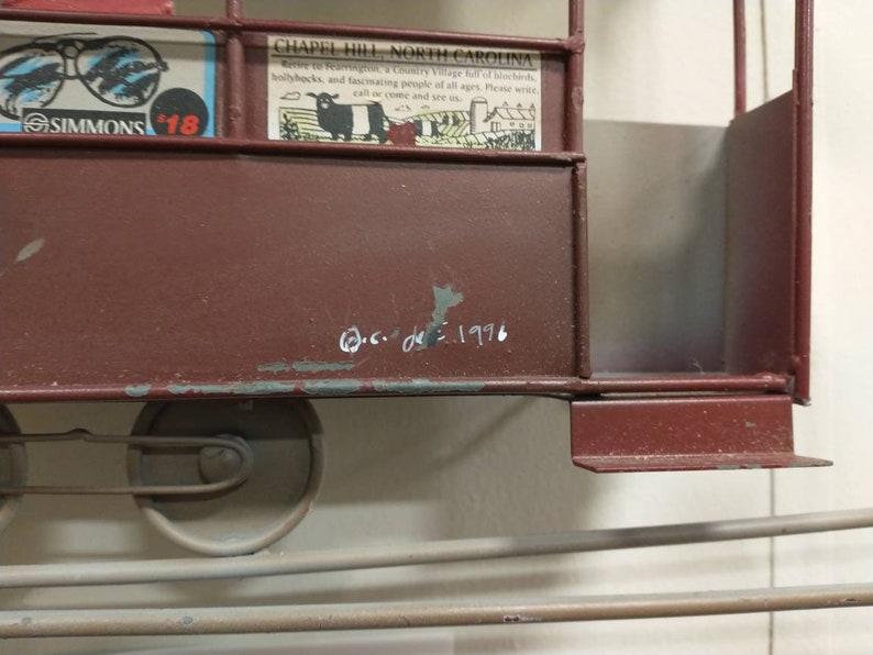 May include: A red metal model of a train car with a white inscription that reads "c.c. de 1996". The train car has a metal frame and a red body. The train car has two wheels on the bottom.