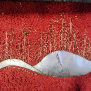 Pode incluir: Uma pe&ccedil;a de arte de parede em metal que retrata uma paisagem de inverno. A obra apresenta &aacute;rvores estilizadas em dourado e uma forma curva prateada, possivelmente uma colina coberta de neve ou um corpo de &aacute;gua. O fundo &eacute; vermelho vibrante.
