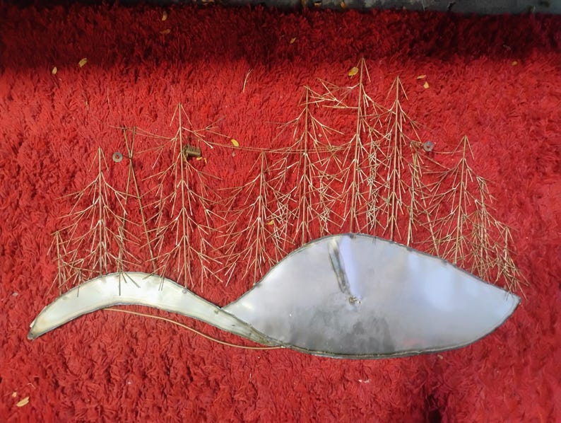 Pode incluir: Uma pe&ccedil;a de arte de parede em metal que retrata uma paisagem de inverno. A obra apresenta &aacute;rvores estilizadas em dourado e uma forma curva prateada, possivelmente uma colina coberta de neve ou um corpo de &aacute;gua. O fundo &eacute; vermelho vibrante.