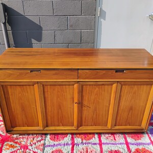 Op de afbeelding: Een rechthoekig houten dressoir met een glad bovenoppervlak, twee horizontale lades met verzonken handgrepen en vier onderste kastdeuren met verticale houtnerfpanelen in een middelbruine houtkleur.