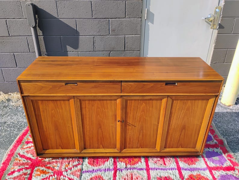 Op de afbeelding: Een rechthoekig houten dressoir met een glad bovenoppervlak, twee horizontale lades met verzonken handgrepen en vier onderste kastdeuren met verticale houtnerfpanelen in een middelbruine houtkleur.