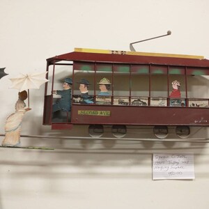 May include: A vintage metal toy trolley car with passengers, hanging from a wire, with two people walking on a wire below. The trolley car is red with a yellow roof and says "Second Ave" on the side. The trolley car is hanging from a wire and is attached to a wall. The price tag says "$95.00".