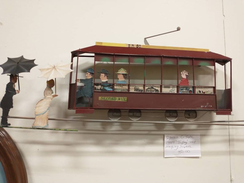 May include: A vintage metal toy trolley car with passengers, hanging from a wire, with two people walking on a wire below. The trolley car is red with a yellow roof and says "Second Ave" on the side. The trolley car is hanging from a wire and is attached to a wall. The price tag says "$95.00".