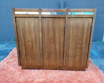 Könnte beinhalten: Ein dreitüriger Holzschrank im Mid-Century-Modern-Stil. Der Schrank hat eine hellbraune Holzoptik und silberne Metallakzente. Jede Tür hat einen horizontalen silbernen Metallstreifen mit einem kleinen silbernen Metallgriff.