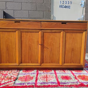 Op de afbeelding: Een rechthoekig houten dressoir met twee horizontale lades aan de bovenzijde met verzonken grepen en vier verticale kastdeuren eronder. Het meubel is vervaardigd uit middelbruin hout met een zichtbare houtnerf en een gladde afwerking.