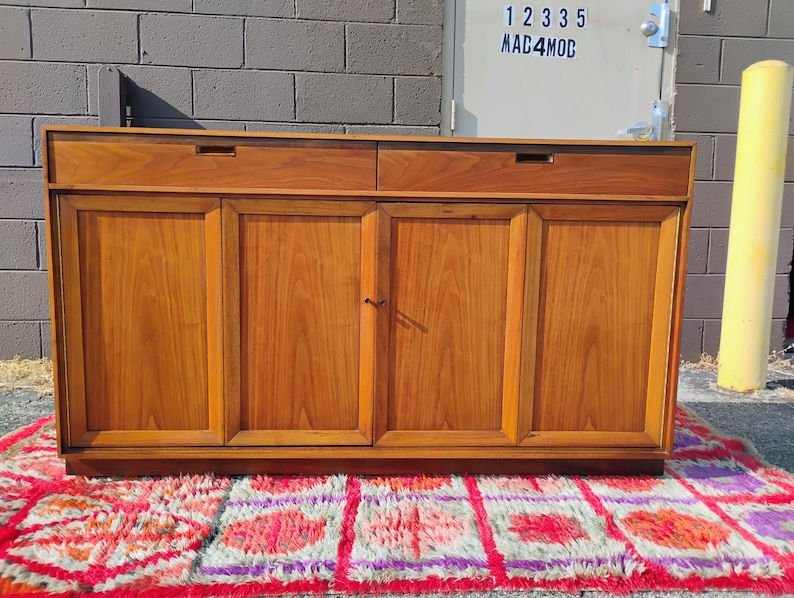 Op de afbeelding: Een rechthoekig houten dressoir met twee horizontale lades aan de bovenzijde met verzonken grepen en vier verticale kastdeuren eronder. Het meubel is vervaardigd uit middelbruin hout met een zichtbare houtnerf en een gladde afwerking.