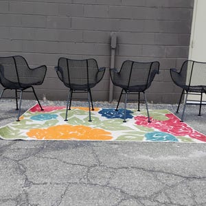 May include: Four black metal chairs with a wire mesh design are arranged in a row on a colorful rug with floral patterns. The chairs have a simple, mid-century modern style.