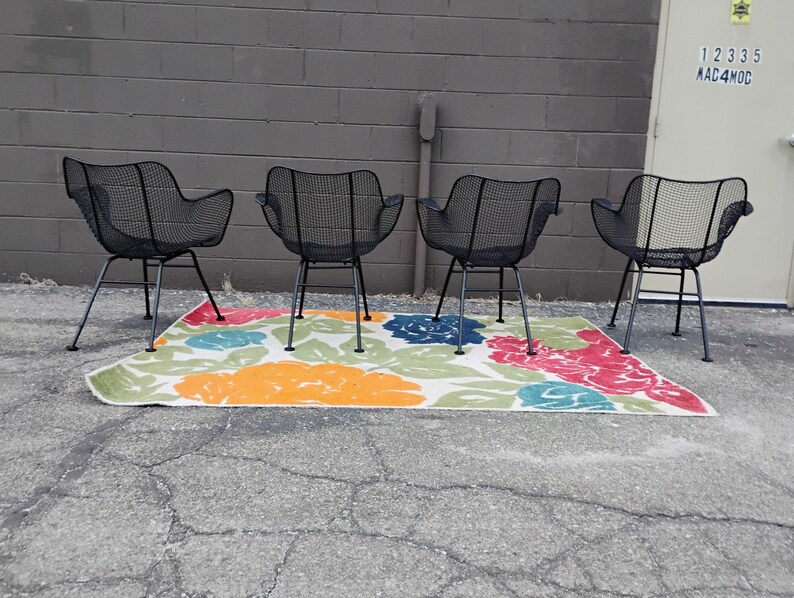 May include: Four black metal chairs with a wire mesh design are arranged in a row on a colorful rug with floral patterns. The chairs have a simple, mid-century modern style.