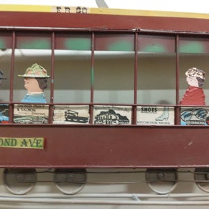 May include: A red and brown metal model of a streetcar with a green and white interior. The streetcar has a sign that reads "Second Ave" and has several small advertisements on the side. The streetcar has four wheels and is on a track.